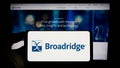Person holding cellphone with logo of US company Broadridge Financial Solutions Inc. on screen in front of webpage.
