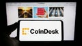 Person holding cellphone with logo of US crypto news company CoinDesk Inc. on screen in front of business webpage. Royalty Free Stock Photo