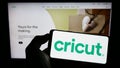 Person holding cellphone with logo of US cutting plotter company Cricut Inc. on screen in front of business webpage.