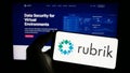 Person holding cellphone with logo of US data security company Rubrik Inc. on screen in front of business webpage.