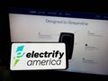 Person holding cellphone with logo of US EV charging company Electrify America LLC on screen in front of business web page.
