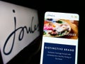 Person holding cellphone with webpage of British retail company Joules Group plc on screen in front of logo.