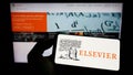 Person holding mobile phone with logo of academic publishing company Elsevier on screen in front of business web page.