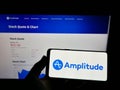 Person holding mobile phone with logo of American analytics software company Amplitude Inc. on screen in front of web page.