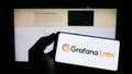 Person holding mobile phone with logo of American analytics software company Grafana Labs on screen in front of web page.