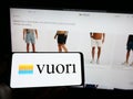 Person holding mobile phone with logo of American apparel e-commerce company Vuori Inc. on screen in front of web page.