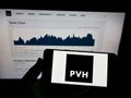 Person holding mobile phone with logo of American clothing company PVH Corp. on screen in front of business web page.