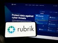 Person holding mobile phone with logo of American data security company Rubrik Inc. on screen in front of web page.