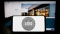 Person holding mobile phone with logo of American green building certification LEED on screen in front of web page.