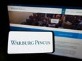 Person holding mobile phone with logo of American private equity company Warburg Pincus LLC on screen in front of webpage.