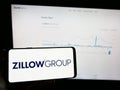 Person holding mobile phone with logo of American real estate company Zillow Group Inc. on screen in front of web page.