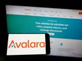 Person holding mobile phone with logo of American software company Avalara Inc. on screen in front of business web page.