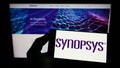 Person holding mobile phone with logo of American technology company Synopsys Inc. on screen in front of web page.