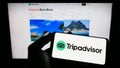 Person holding mobile phone with logo of American travel company Tripadvisor Inc. on screen in front of business web page.