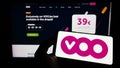 Person holding mobile phone with logo of Belgian telecommunications company Voo SA on screen in front of business web page.