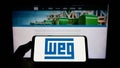 Person holding mobile phone with logo of Brazilian company WEG Equipamentos Eletricos S.A. on screen in front of web page.