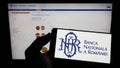 Person holding mobile phone with logo of central bank Banca Nationala a Romaniei (BNR) on screen in front of web page.