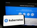Person holding mobile phone with logo of container orchestration system Kubernetes on screen in front of web page.
