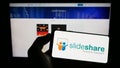 Person holding mobile phone with logo of content hosting company SlideShare on screen in front of business web page.