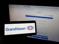 Person holding mobile phone with logo of Dutch optical retailing company GrandVision N.V. on screen in front of web page.