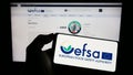 Person holding mobile phone with logo of EU agency European Food Safety Authority (EFSA) on screen in front of web page. Royalty Free Stock Photo