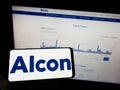 Person holding mobile phone with logo of eye care company Alcon AG on screen in front of business web page.