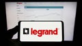 Person holding mobile phone with logo of French electronics company Legrand SA on screen in front of business web page.