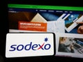 Person holding mobile phone with logo of French facility management company Sodexo S.A. on screen in front of web page.