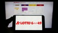 Person holding mobile phone with logo of German lottery Deutsche Lotto- und Totoblock on screen in front of web page.