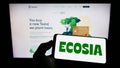 Person holding mobile phone with logo of German search engine company Ecosia GmbH on screen in front of business web page.