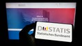 Person holding mobile phone with logo of German Statistisches Bundesamt (Destatis) on screen in front of web page.