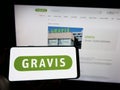 Person holding mobile phone with logo of Gravis Computervertriebsgesellschaft mbH on screen in front of web page.