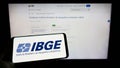 Person holding mobile phone with logo of Instituto Brasileiro de Geografia e Estatistica (IBGE) on screen with web page.