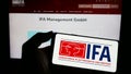 Person holding mobile phone with logo of Internationale Funkausstellung Berlin (IFA) on screen in front of web page.