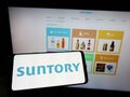 Person holding mobile phone with logo of Japanese beverage company Suntory Holdings KK on screen in front of web page.