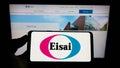 Person holding mobile phone with logo of Japanese pharmaceutical company Eisai Co. Ltd. on screen in front of web page.