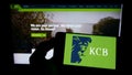 Person holding mobile phone with logo of Kenyan financial company KCB Group Limited on screen in front of web page.