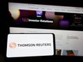 Person holding mobile phone with logo of media company Thomson Reuters Corporation on screen in front of business webpage.