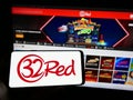 Person holding mobile phone with logo of online casino company 32Red Limited on screen in front of business web page.
