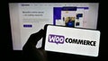 Person holding mobile phone with logo of open-source e-commerce solution WooCommerce on screen in front of web page.