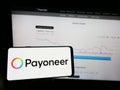 Person holding mobile phone with logo of payments company Payoneer Global Inc. on screen in front of business web page.