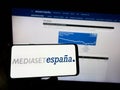 Person holding mobile phone with logo of Spanish media company Mediaset Espana on screen in front of business web page.