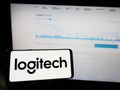 Person holding mobile phone with logo of Swiss company Logitech International SA on screen in front of business web page.