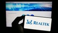 Person holding mobile phone with logo of Taiwanese company Realtek Semiconductor Corp. on screen in front of web page. Royalty Free Stock Photo