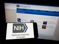 Person holding mobile phone with logo of US agency National Institutes of Health (NIH) on screen in front of web page.