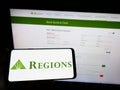 Person holding mobile phone with logo of US banking company Regions Financial Corporation on screen in front of web page.