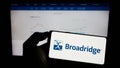 Person holding mobile phone with logo of US company Broadridge Financial Solutions Inc. on screen in front of web page.