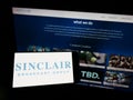 Person holding mobile phone with logo of US company Sinclair Broadcast Group Inc. (SBG) on screen in front of web page.