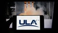 Person holding mobile phone with logo of US space company United Launch Alliance (ULA) on screen in front of web page.