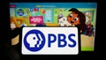 Person holding mobile phone with logo of US tv network Public Broadcasting Service (PBS) on screen in front of web page.